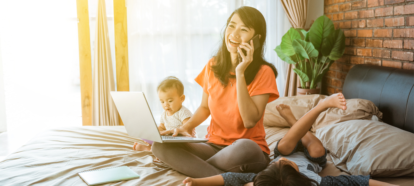 Mom on her bed with two children with a laptop in her lap and a cellphone in her hand.