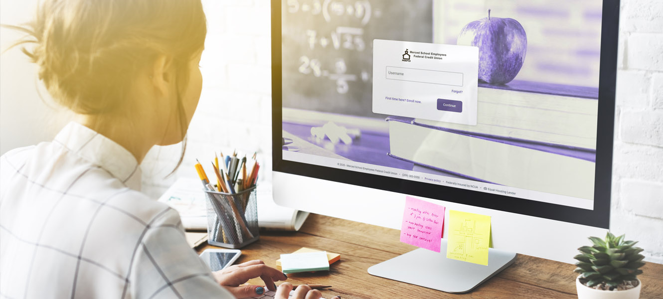 A woman sitting on her desktop computer looking at the screen. The wesbite on the screen is for cu@home online banking.