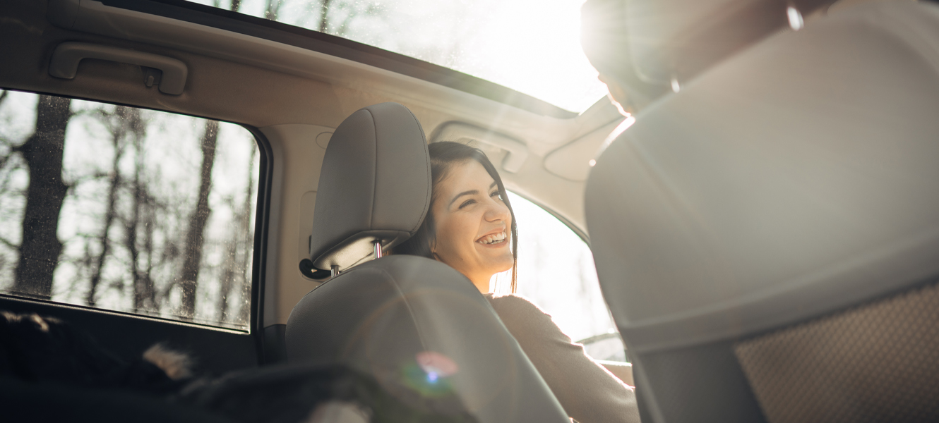 A woman sitting in the front seat of her vehicle looking over and smiling at someone in the passenger seat.