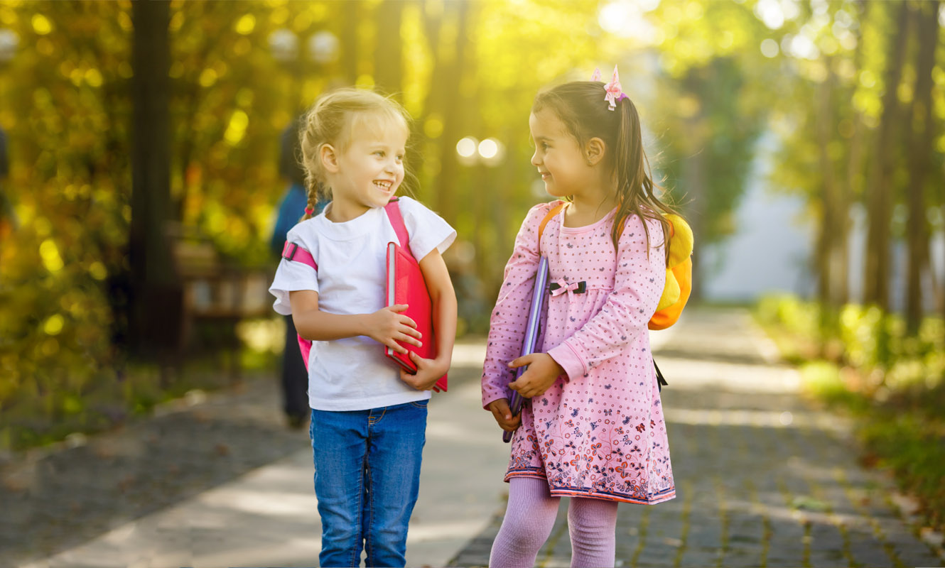 Two little girls with backpacks on walking down the side walk looking at eachother.